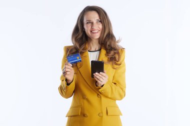 Bu orta boy fotoğraf sarı ceketli gülümseyen profesyonel bir kadını gösteriyor. Elinde kredi kartı ve akıllı telefon var. Dijital finansal işlemler ve modern ticareti somutlaştırır..