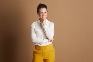 Beyaz gömlekli ve sarı pantolonlu kendine güvenen bir iş kadını eli çenesinde özenle poz veriyor, düz kahverengi arka planda kameraya gülümsüyor..