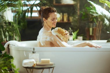 Gözleri kapalı, gülümseyen, mutlu bir köpük banyosundan zevk alan, omzunu doğal bir süngerle ovuşturan bir kadın. Etrafı yemyeşil bitkilerle çevrili olan sahne, dingin bir rahatlama yayar..