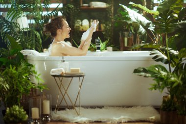 Çörekli neşeli bir kadın modern beyaz köpük banyosunda neşeyle gülüyor, elleri köpükle kaplı, bol yeşil bitkilerle çevrili, lüks ve huzurlu bir spa merkezi yaratıyor..