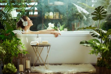 Gözleri kapalı bir kadın, yemyeşil bitkilerin ve yumuşak sisin ortasında kulaklık takarak beyaz bir küvette dinleniyor. Bu resim, spa benzeri bir ev ortamında derin bir sükunet ve kendine bakım yakalar.