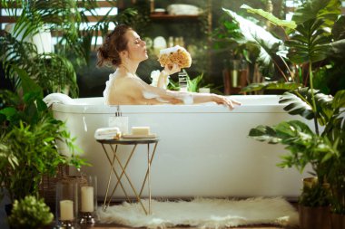 Gözleri kapalı sakin bir kadın, elinde doğal bir deniz süngeri olan, beyaz bir küvette rahatlatıcı köpük banyosundan zevk alır. Bereketli yeşil bitkilerle çevrili ve yumuşak bir ışık yayar, sakin, kaplıca benzeri bir atmosfer..