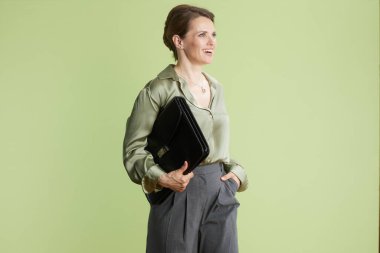 Açık yeşil saten gömlekli ve gri pantolonlu orta yaşlı bir kadın açık yeşil bir fona karşı elinde siyah bir evrak çantası tutarken gülümsüyor. Profesyonel zarafetin bir resmi..