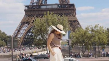 Yazlık şapkalı ve şık beyaz pantolonlu kendine güvenen bir kadın Eyfel Kulesi 'nin önünde gülümsüyor ve Paris' te aydınlık bir günün tadını çıkarıyor..