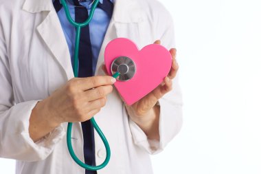Beyaz önlüklü bir doktorun, kalp şeklindeki pembe bir objeyi incelemek için steteskop kullanması kardiyovasküler sağlık ve tıbbi bakımı temsil ediyor..