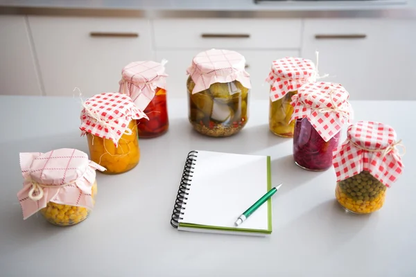 Jars of preserved vegetables on kitchen counter with notepad — Stock Photo, Image