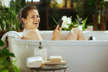 Açık kahverengi saçlı yetişkin bir kadın köpüklü beyaz küvette gülüyor, elinde akıllı telefon ve kameraya bakıyor. Yemyeşil bitkiler huzur, doğal ortam, mutluluk ve rahatlama yaratır..