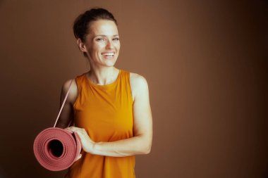 30 'lu yaşlarda, hardal sarısı kolsuz bluz giyen neşeli bir kadın pembe kayışlı yoga minderini tutarken gülümsüyor. Kapalı bir stüdyo ortamında çalışmaya hazır..