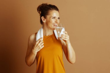 Portakal rengi bluzlu gülümseyen orta yaşlı bir kadın boynunda beyaz bir havluyla bardaktan su içiyor. Tatbikat sonrası memnuniyetini ve refahını ifade etmek için yana bakıyor..