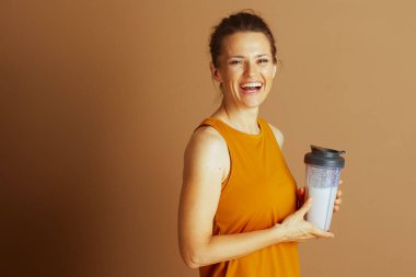 Sarı kolsuz tişörtlü neşeli bir kadın gülümsüyor, elinde temiz protein içeceği var. Açık kahverengi arka planda canlı, aktif ve sağlıklı bir yaşam tarzına sahip, sağlığını ve günlük rutinini ön plana çıkarıyor.