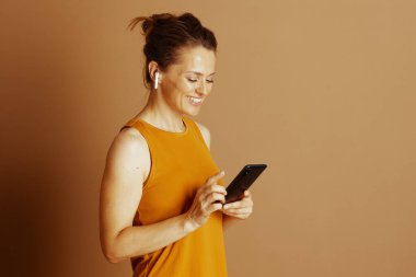 Sarı bluzlu rahat bir kadın, kulaklık takıyor, akıllı telefonuna gülümsüyor. Bu resim modern teknoloji kullanımını, dijital bağlantıyı ve gelişigüzel, bağlı bir yaşam tarzını resmediyor..