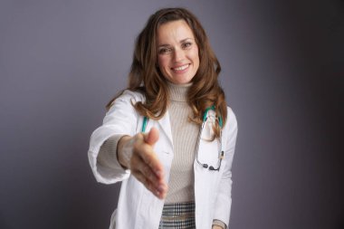 Beyaz laboratuvar önlüklü profesyonel bir kadın doktor elini uzatıyor. Karşılayıcı bir gülümsemeyle, güveni, tıbbi muayeneyi ve hasta bakımını temsil ediyor..