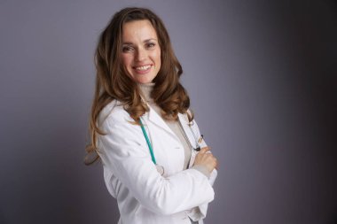 Kollarını kavuşturmuş, beyaz laboratuvar önlüğü ve steteskop takmış, uzmanlık ve güvenilir sağlık hizmetleri sunan kendine güvenen bir kadın doktor duruyor..