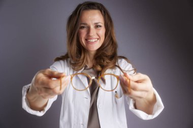 Beyaz laboratuvar önlüklü, dost canlısı bir bayan doktor, görüş bakımı, optometri ve profesyonel göz muayenelerini temsil eden bir çift şık kahverengi gözlük tutuyor..