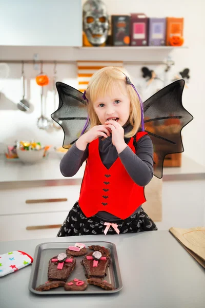 Chica rubia en traje de murciélago comiendo galletas de Halloween en la cocina — Foto de Stock