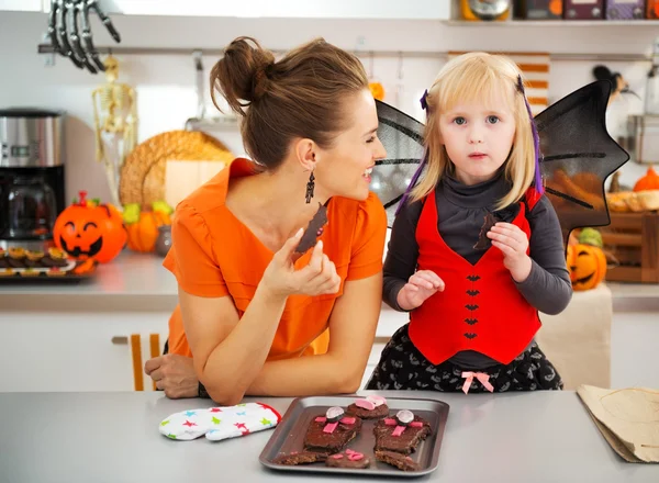 Chica en traje de murciélago con la madre sonriente comiendo galletas de Halloween —  Fotos de Stock