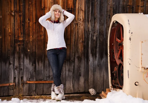 Relaxed young woman in furry hat standing near rustic wood wall — Stockfoto