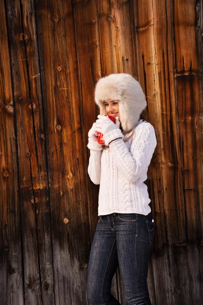 Glückliche Frau mit Pelzmütze und roter Tasse an rustikaler Holzwand — Stockfoto