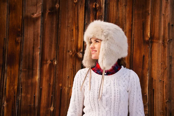 Happy woman in furry hat near rustic wood wall looking aside — Stockfoto