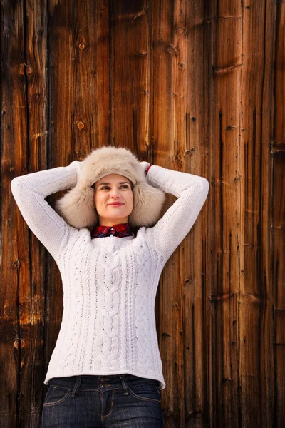 Mujer relajada en suéter de punto y sombrero peludo cerca de la pared de madera —  Fotos de Stock