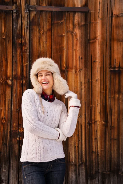 Happy woman in white sweater and furry hat near rustic wood wall — Stock fotografie