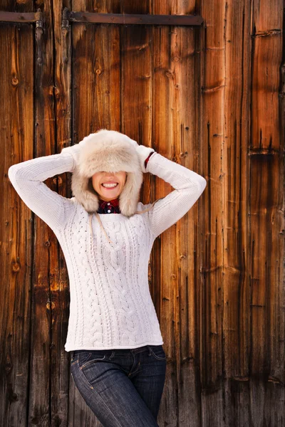 Young woman pulled furry hat over her eyes near rustic wood wall — 图库照片