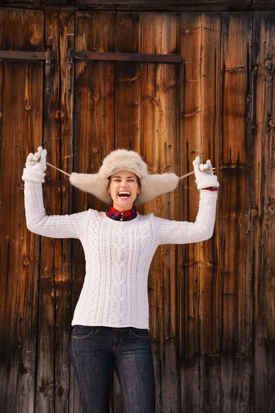 Mujer sonriente jugueteando con sombrero peludo cerca de la pared de madera —  Fotos de Stock