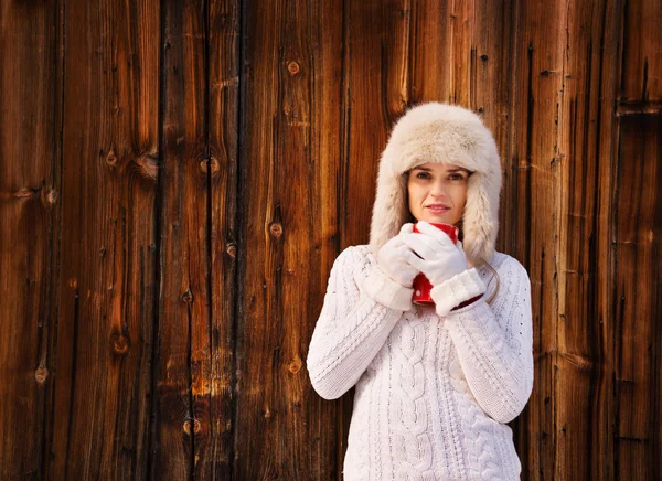 Женщина в белом трикотажном свитере с чашкой возле деревенской деревянной стены — стоковое фото