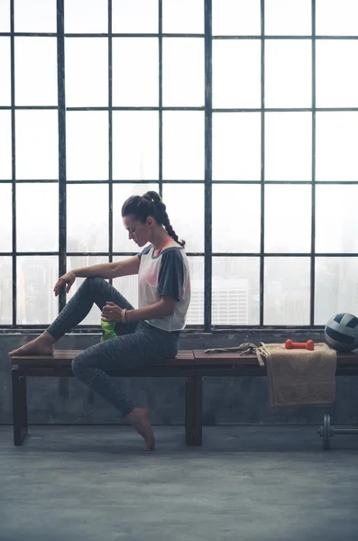 Fit woman sitting on bench in loft gym, looking at water bottle — Stock Photo, Image