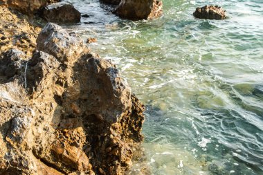 Kristal berrak Akdeniz suları, parlak bir yaz güneşi altında engebeli deniz taşlarını nazikçe yıkıyor.