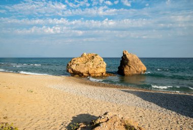 Sakin kıyı manzarası, büyük sahil kayaları, yumuşak dalgalar, parlak kum ve berrak mavi su ile huzurlu bir gökyüzü altında.