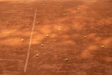 Boş bir kil kortunda çoklu tenis topu uzanır. Bu da antrenman yoğunluğunu ve spor hazırlıklarını yansıtır.