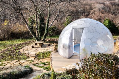 Açık havada jakuziyi kaplayan şeffaf polikarbonat kubbesi dağ villa bahçesinde, güneşli kış manzarasında modern spa barınağı