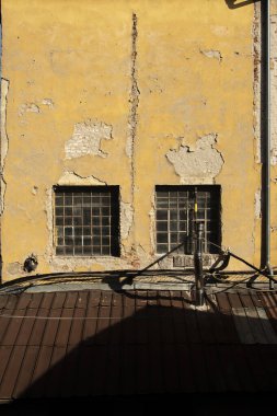 Çatlak alçılı, sarı boyalı ve metal parmaklıklı camları olan eski, bakımsız bir endüstriyel binanın arka cephesi. Yıpranmış dokuları ve endüstriyel atmosferi olan kentsel çürüme mimarisi.