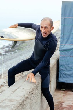 Ellerinde sörf tahtasıyla sahilde duran olgun sörfçü portresi..