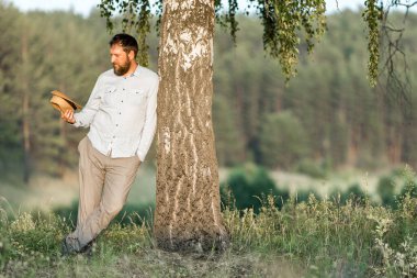 Yetişkin bir adam ağaca yaslanıyor ve kafasından çıkardığı şapkayı izliyor. Ağacın yanındaki sakin adam.