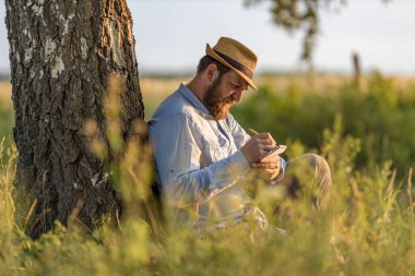 Sakallı yetişkin bir adam şiir yazar ya da bir ağacın yanında yazı yazar. Doğa yazma kavramı
