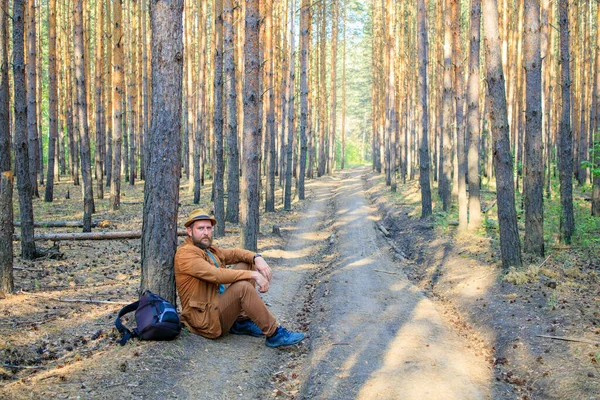 a tired man sat down to rest near a tree on a forest road, a bearded man in a hat. traveler in recovery