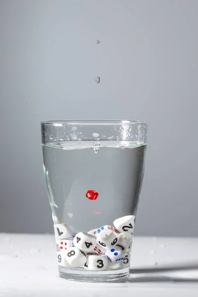 in a glass of water at the bottom are cubes with white numbers, in the middle of the glass is a red cube. a drop of water over a glass