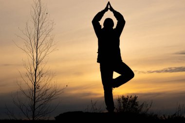 Bir adamın silueti, birden fazla bacağıyla ayakta dururken kollarını başının üzerine katlayarak meditasyon yapan bir adamın silüeti. günbatımında gökyüzü