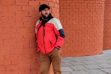 Sakallı bir adam sırtı tuğla bir duvara dönük durur ve bakar. Eller pantolonun cebinde.