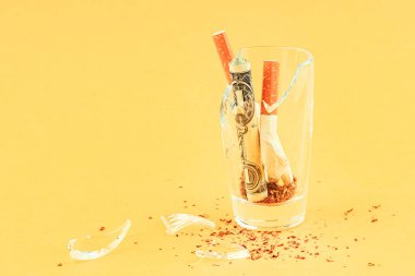 Kırık sigara ve kırık bardakta yuvarlanmış bir dolar. sarı arkaplan bir tütün serpintisi