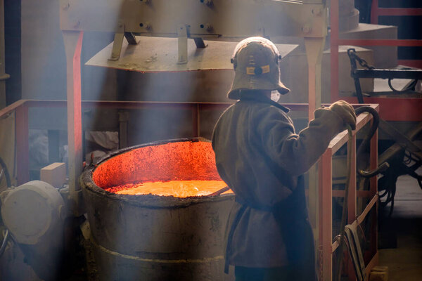 a worker with a metal pole at a vat of molten red metal. foundry job