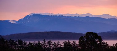 Pankart için yatay bir fotoğraf düzenleme. Dağlarda şafak, ormanın içinden görünür. Sabahın erken saatlerinde dağların karlı zirvelerini görmek