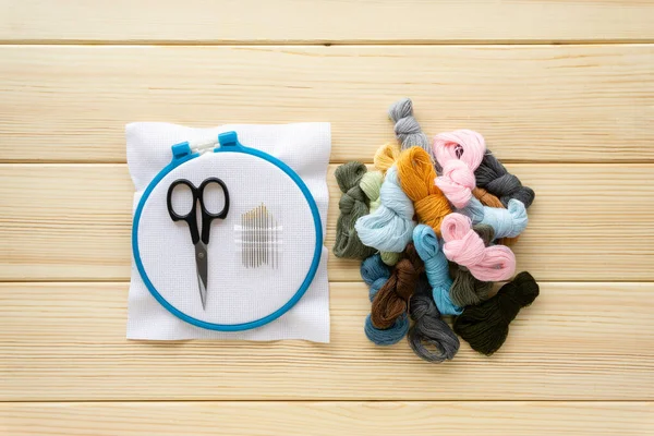 Flat lay of items for embroidery. Hoop, floss threads, needles and canvas on a wooden background