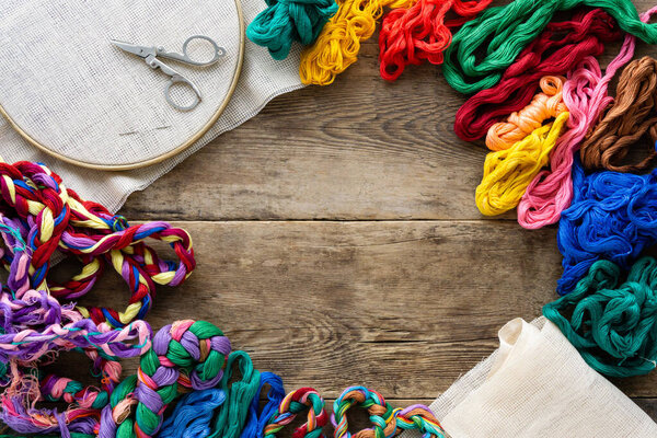 Frame of accessories for embroidery. Hoop, colorful floss threads and canvas on old wooden background, copyspace