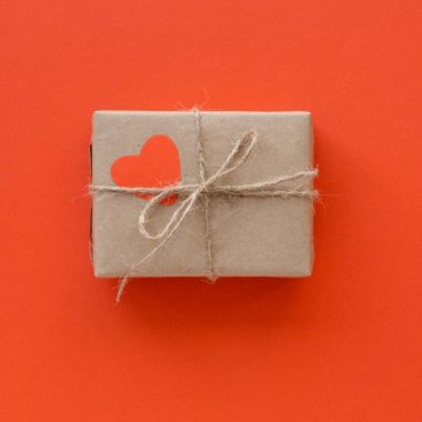 Kırmızı arka planda dekoratif kalbi olan el sanatları kağıdına sarılı bir hediye kutusu.