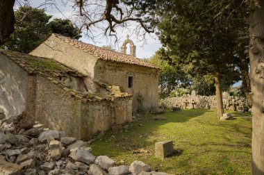 Cres, Hırvatistan - 9 Nisan 2023: Cres Hırvatistan 'da baharda bulutlu gökyüzü altında mezarlığı ve ağaçları olan küçük bir taş şapel bulunuyor.