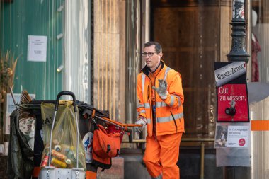 Viyana, Avusturya - 7 Nisan 2020: Turuncu üniformalı bir atık toplayıcı baharda sokakta çöp kamyonunun yanında yürüyor