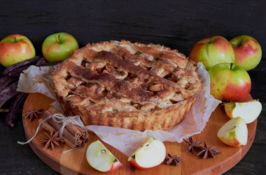Traditional apple pie with spices on the brown wooden board. Apple pie decorated with sliced fresh apples, carob pods and cinnamon sticks  on the black background. Side view 